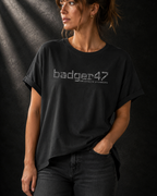 Schwarzes Badger 47 Multicam Grau Damen-T-Shirt mit weitem Schnitt, gerollten Ärmeln, längerem Rückensaum und grauem Camouflage-Logo vor dunklem Studiohintergrund.
