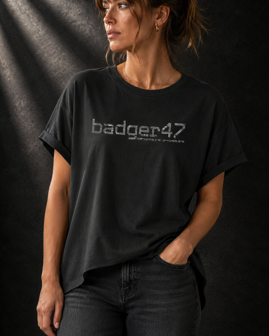 Schwarzes Badger 47 Multicam Grau Damen-T-Shirt mit weitem Schnitt, gerollten Ärmeln, längerem Rückensaum und grauem Camouflage-Logo vor dunklem Studiohintergrund.
