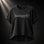 Schwarzes Badger 47 Multicam Grau Damen-T-Shirt mit weitem Schnitt, gerollten Ärmeln, längerem Rückensaum und grauem Camouflage-Logo vor dunklem Studiohintergrund
