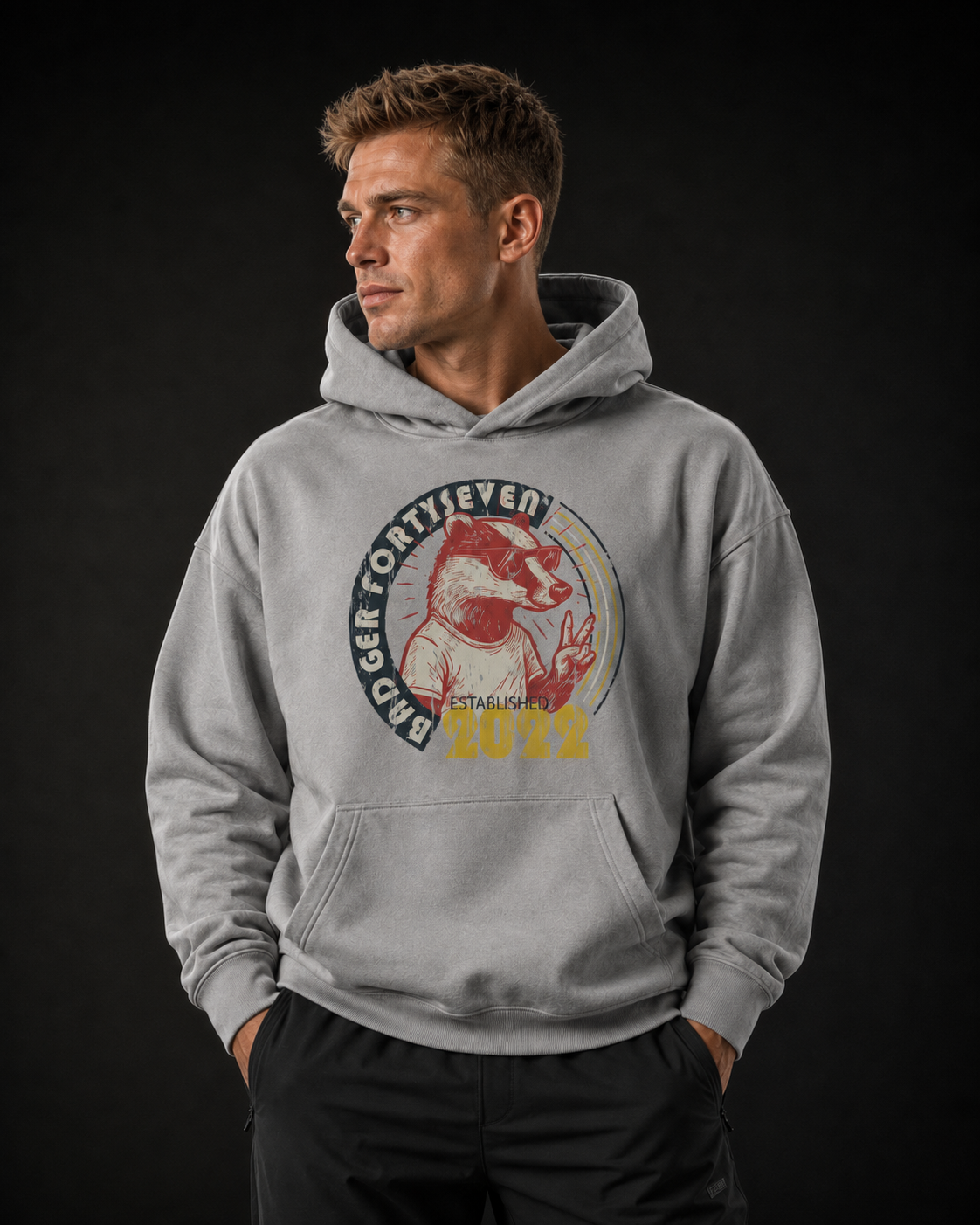 Dunkelblondes männliches Model trägt den Badger Forty Seven Vintage Hoodie in Light Asphalt mit großem Frontprint und schwarzer Stretch-Hose vor dunklem Studiohintergrund.