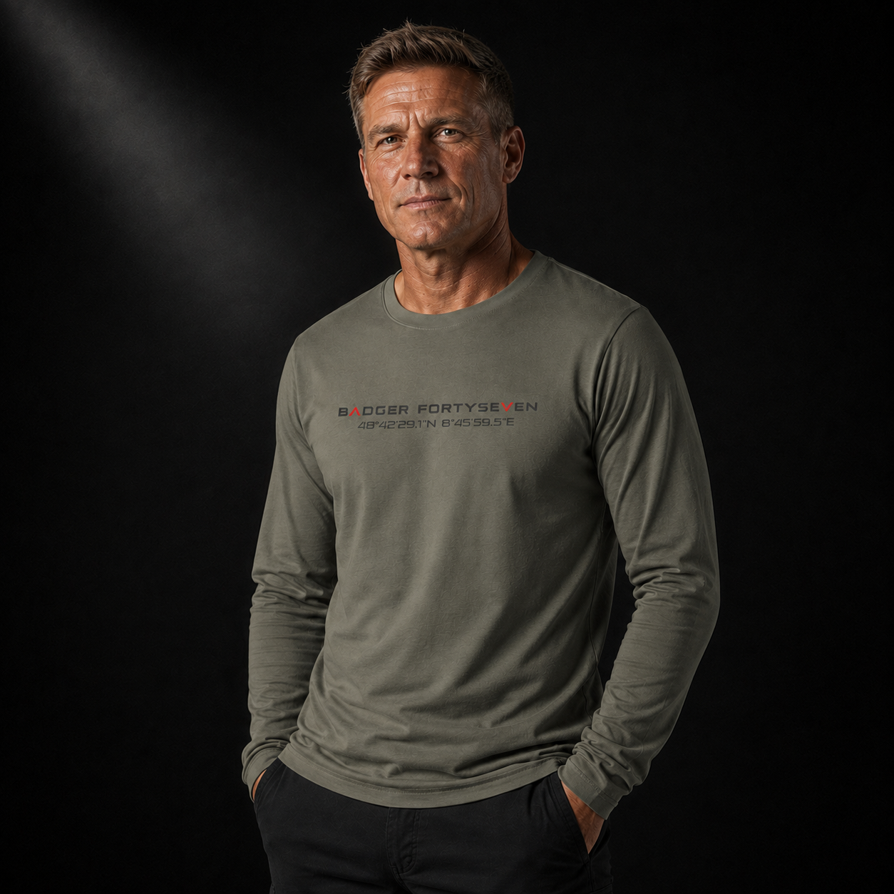 Ca. 50-jähriger männlicher Veteran in khakifarbenem Badger FortySeven Organic Longsleeve mit Koordinaten-Print, leicht gedrehte Pose vor schwarzem Hintergrund mit markantem Studio-Lichteinfall.