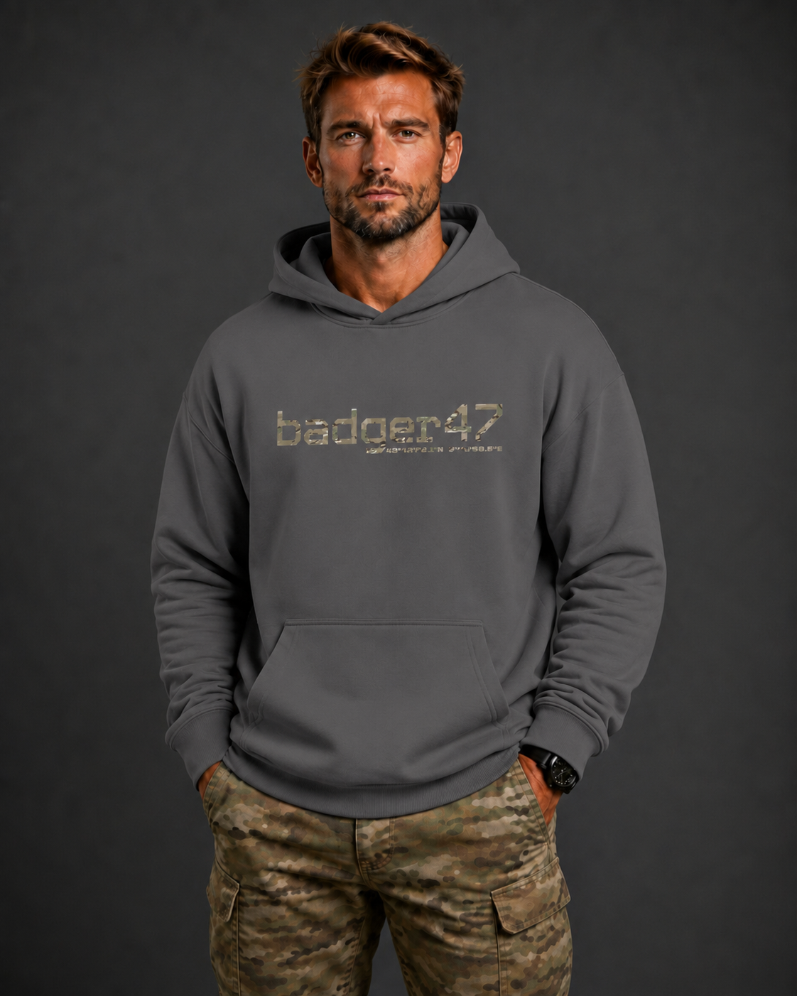 Dunkelblondes Männermodel mit badger47 Boxcut Hoodie in Magnet und Multicam-Hose vor anthrazitfarbenem Studiohintergrund