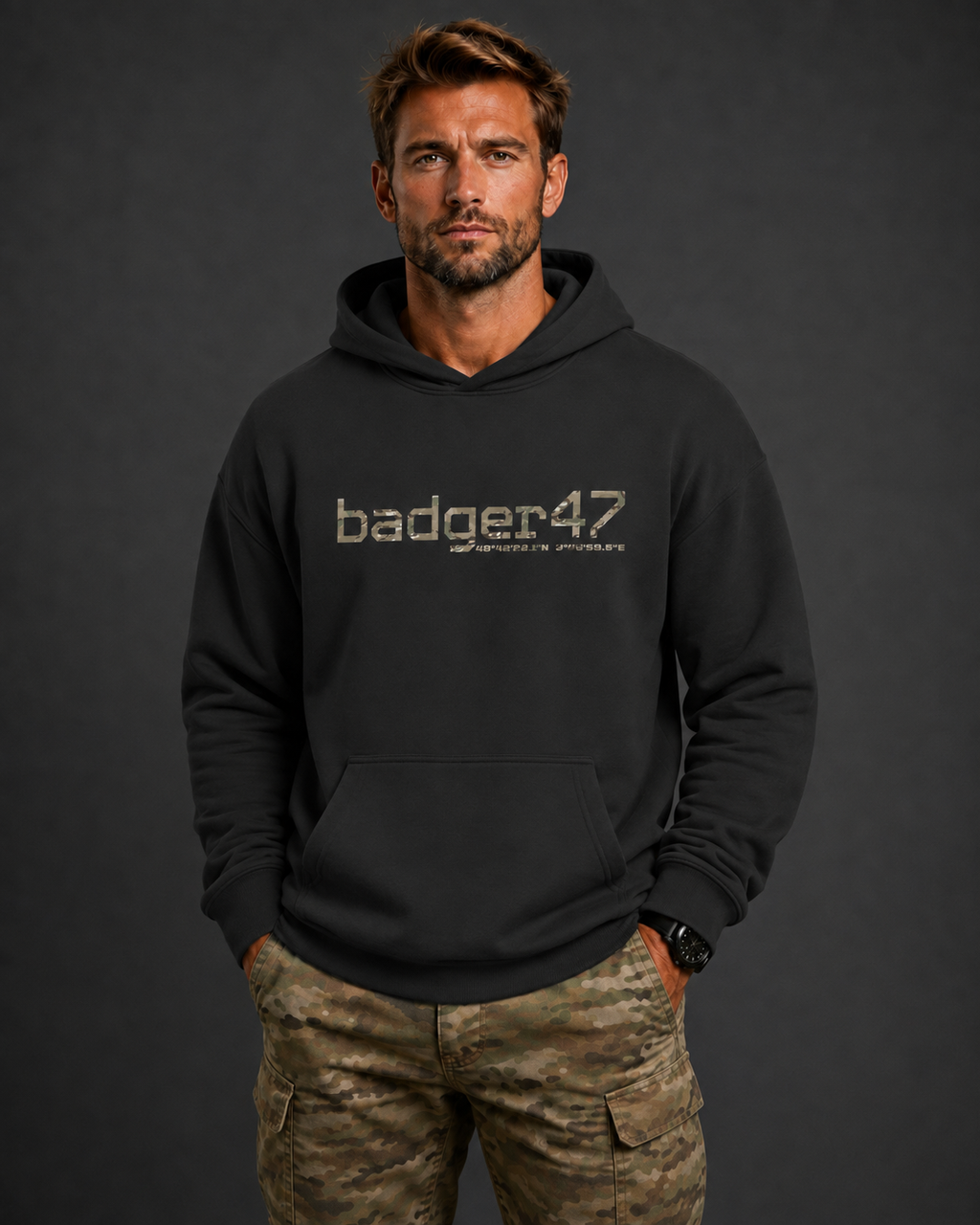 Dunkelblondes Männermodel mit schwarzem badger47 Boxcut Hoodie und Multicam-Hose vor anthrazitfarbenem Studiohintergrund