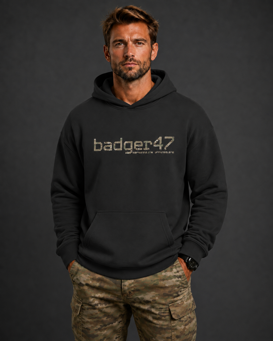 Dunkelblondes Männermodel mit schwarzem badger47 Boxcut Hoodie und Multicam-Hose vor anthrazitfarbenem Studiohintergrund