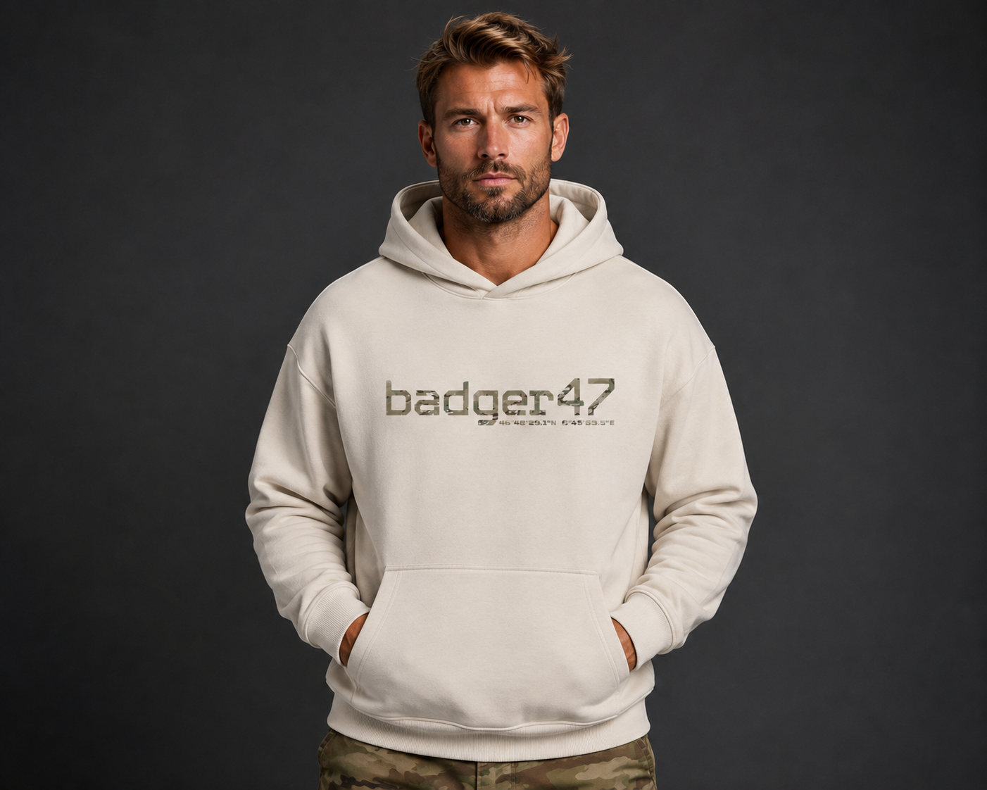 Dunkelblondes Männermodel mit badger47 Boxcut Hoodie in White Sand und Multicam-Hose vor anthrazitfarbenem Studiohintergrund