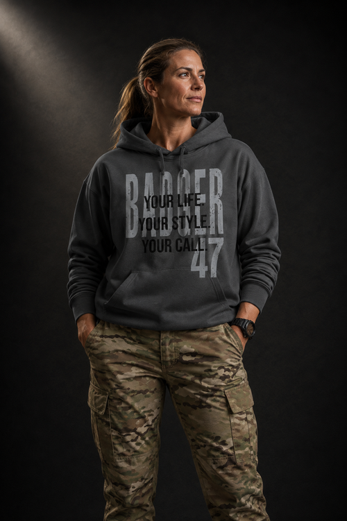 Athletisches Frauenmodel trägt anthrazitfarbenen Badger47 Hoodie mit „Your Life. Your Style. Your Call.“-Print und Multicam-Cargohose vor dunklem Studiohintergrund.