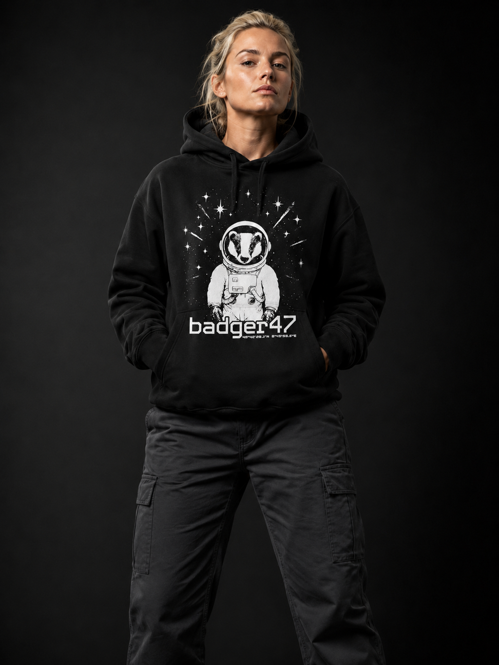 Blondes Frauenmodel Anfang 30 trägt schwarzen Badger47 Hoodie mit Astronauten-Dachs-Print und anthrazitfarbener Utility-Hose vor dunklem Studiohintergrund