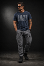 Männliches Model trägt das Badger47 Distressed Organic T-Shirt in India Ink Grey mit grauem BADGER-Frontprint, kombiniert mit grauer Cargo-Hose und schwarzen Boots vor dunklem Studiohintergrund.