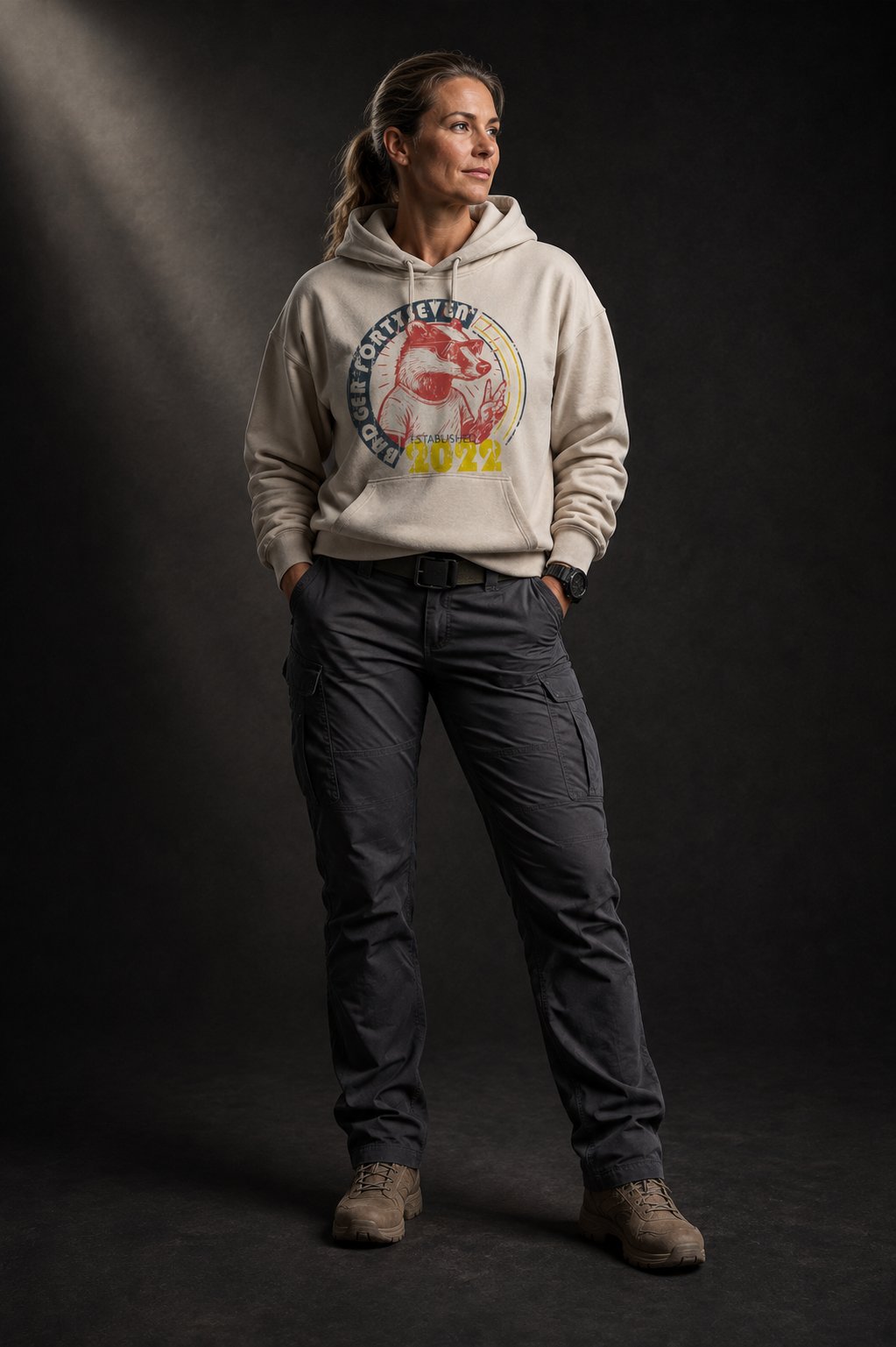 Selbstbewusstes Frauenmodel mit hellbraunen Haaren trägt beigen Badger47 Hoodie mit Vintage-Print und dunkler Utility-Hose vor dunklem Studiohintergrund.