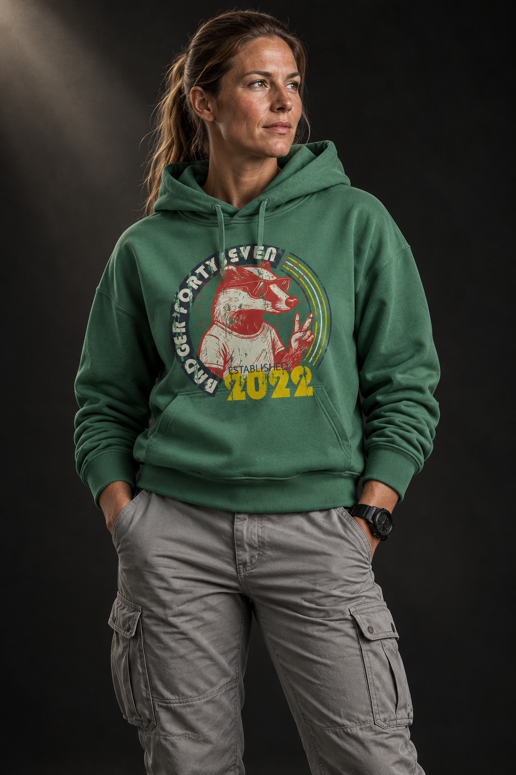 Athletisches Frauenmodel mit hellbraunen Haaren trägt grünen Badger47 Hoodie mit Retro-Print und hellgrauer Utility-Hose vor dunklem Studiohintergrund mit Lichteinfall