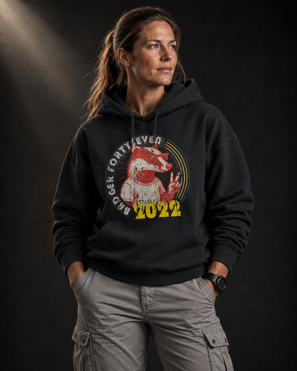 Souveränes Frauenmodel mit hellbraunen Haaren trägt schwarzen Badger47 Hoodie mit Retro-Print und hellgrauer Utility-Hose vor dunklem Studiohintergrund