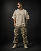 ännliches Model trägt das Badger47 Oversized T-Shirt in Stone mit Multicam-Logo auf der Brust, kombiniert mit Multicam-Cargohose und Boots vor dunklem Studiohintergrund.