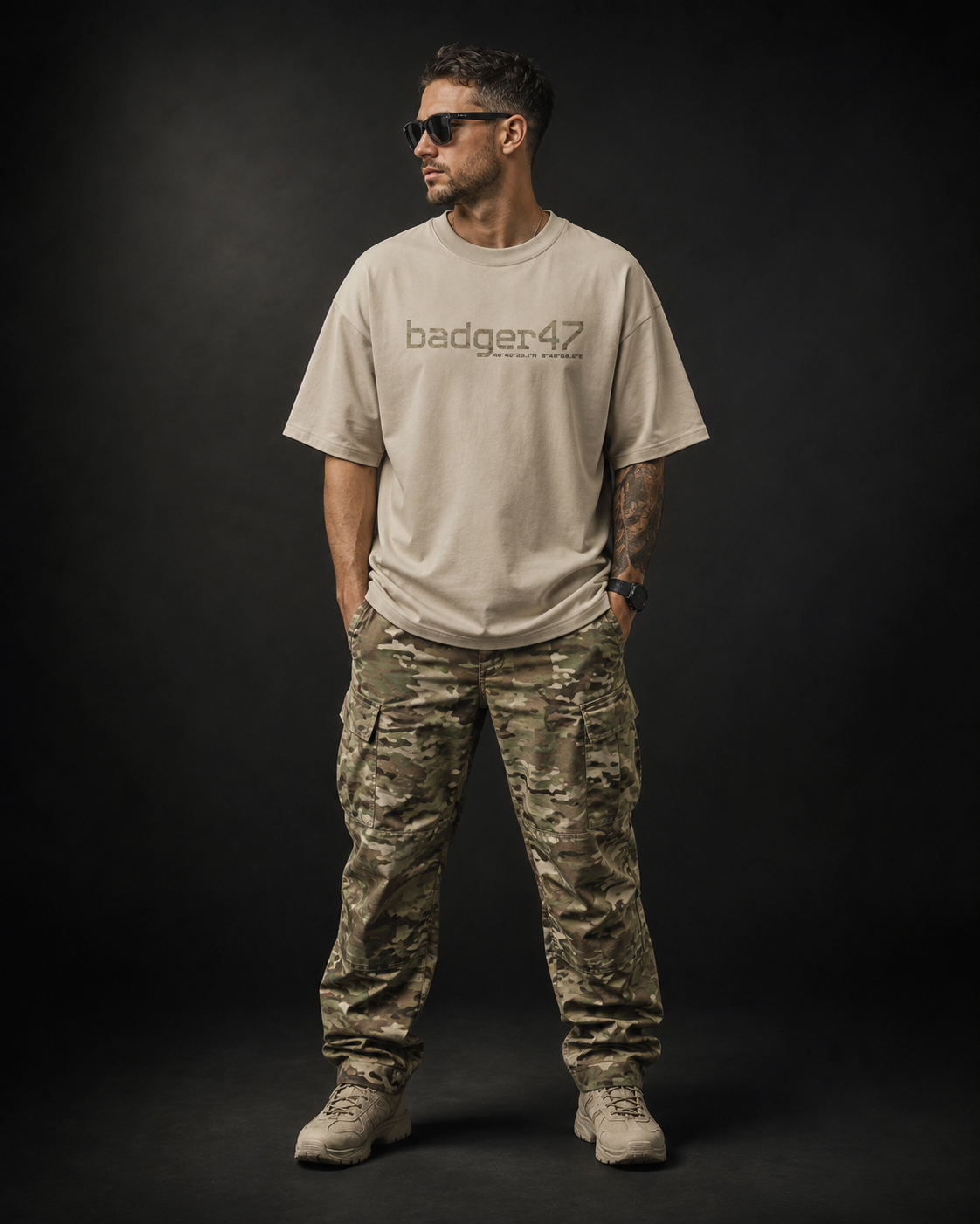 ännliches Model trägt das Badger47 Oversized T-Shirt in Stone mit Multicam-Logo auf der Brust, kombiniert mit Multicam-Cargohose und Boots vor dunklem Studiohintergrund.