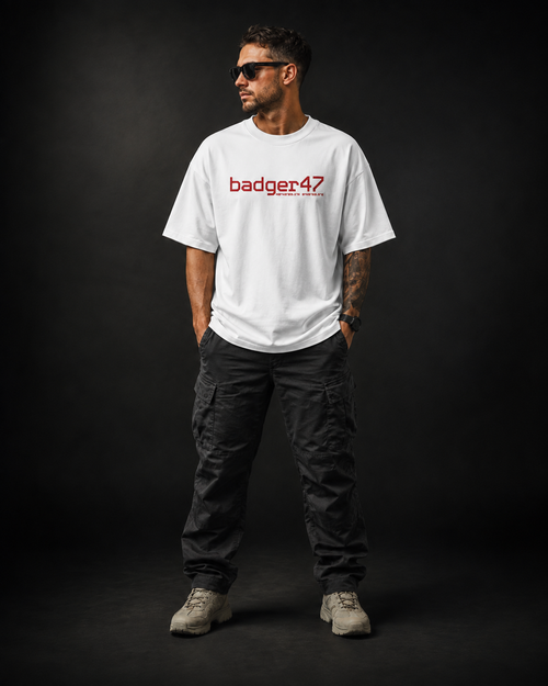 Männliches Model trägt das weiße Badger47 Strive Oversized T-Shirt mit rotem badger47-Frontprint, kombiniert mit schwarzer Cargo-Hose und hellen Sneakers vor dunklem Studiohintergrund.