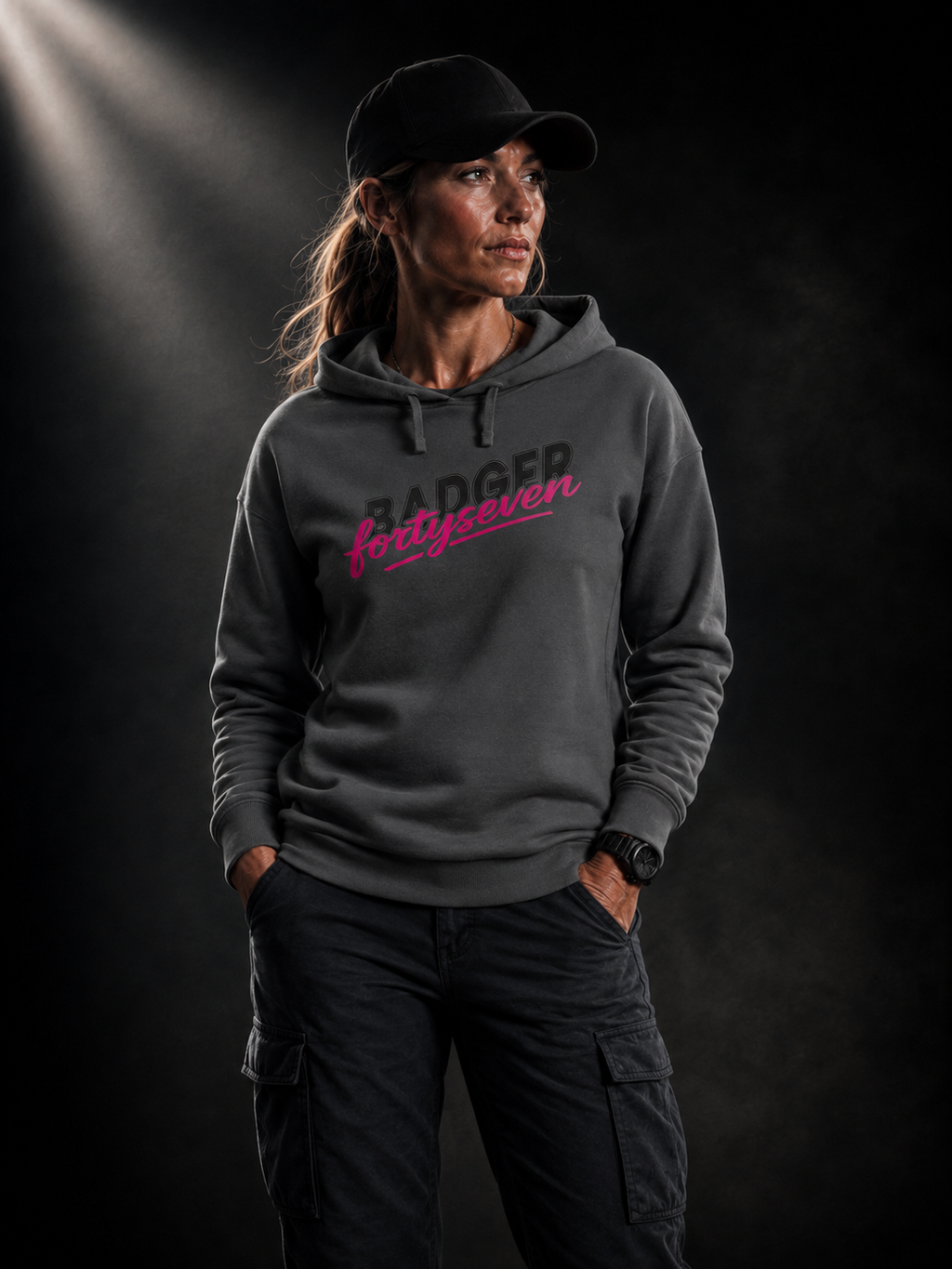 BADGER47 Damen Hoodie in Anthrazit mit BADGER fortyseven Frontprint, getragen von einem Frauenmodel im dunklen Studio mit markantem Lichteinfall von oben links.