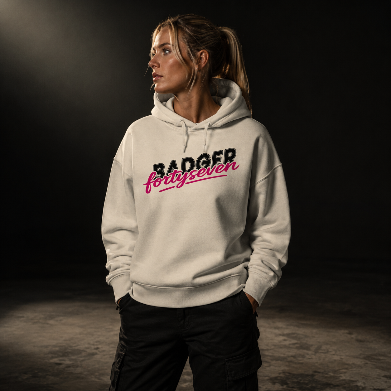 BADGER47 Damen Hoodie in Natural Raw mit BADGER fortyseven Frontprint, getragen von einem Frauenmodel im dunklen Studio mit warmem Lichteinfall von oben links.