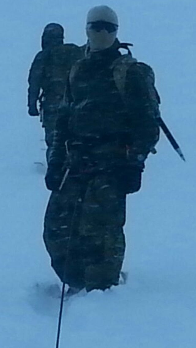 Two operators in winter tactical gear moving through deep snow in a harsh mountain environment at early morning